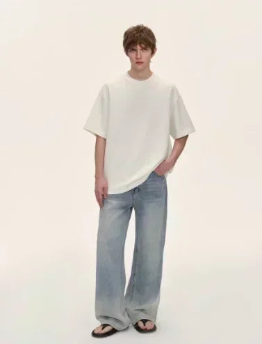 Young man wearing a casual outfit with a white t-shirt and light blue jeans standing against a pale background.